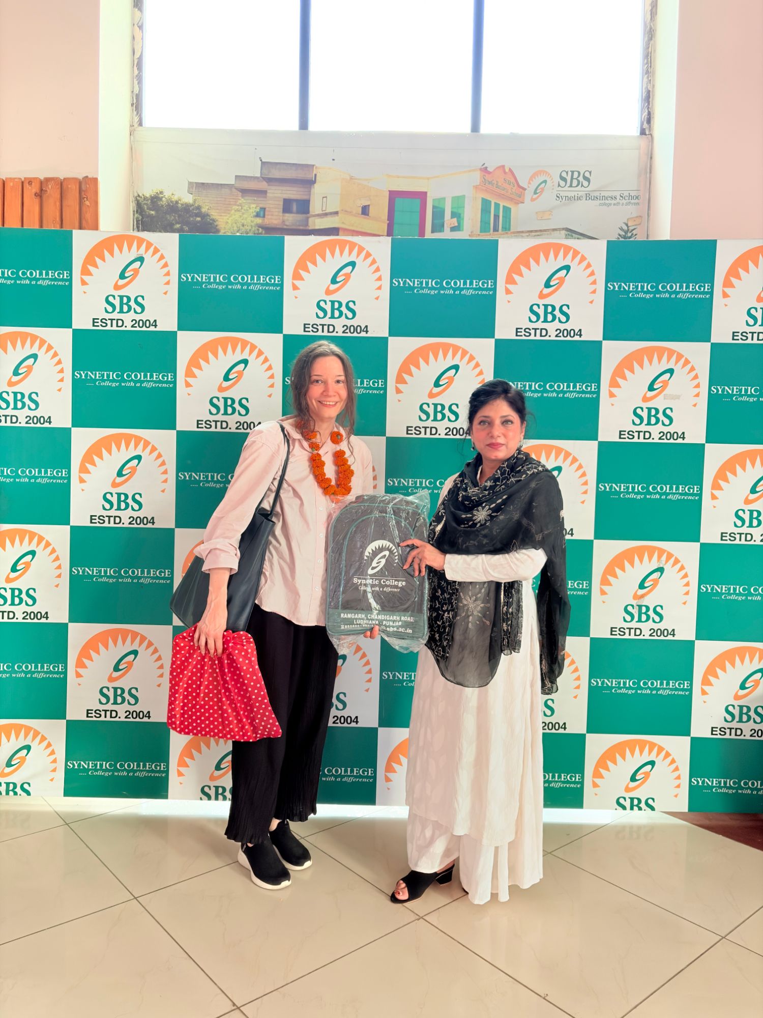 Diana Alexandra Iovino from Finland being welcomed to SBS Synetic College with a faculty member and a Synetic College backpack, in front of the SBS step-and-repeat backdrop