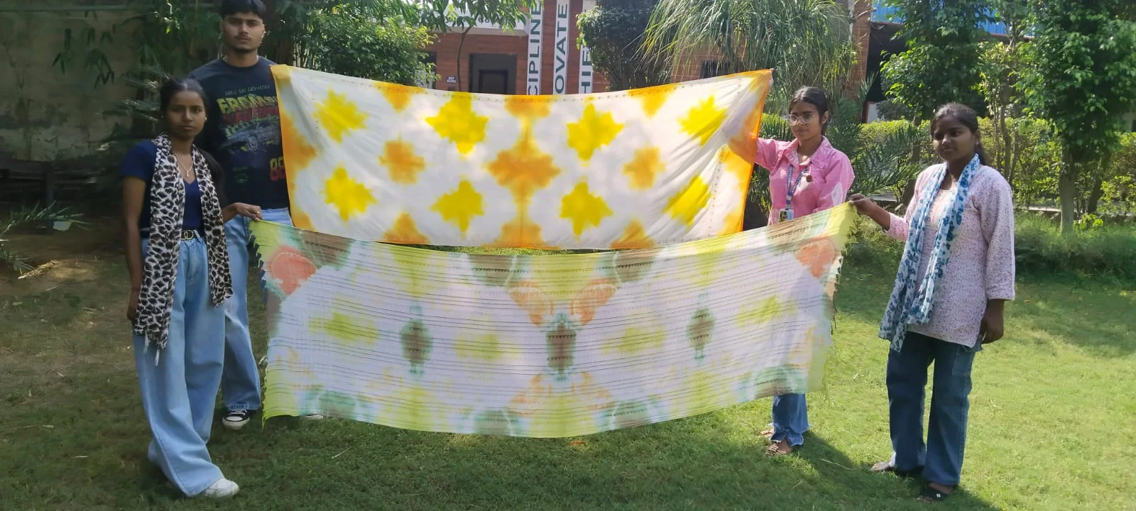 Four B.Sc Textile students holding up two freshly dyed tie-dye fabrics in yellow, orange and green on the SBS campus lawn